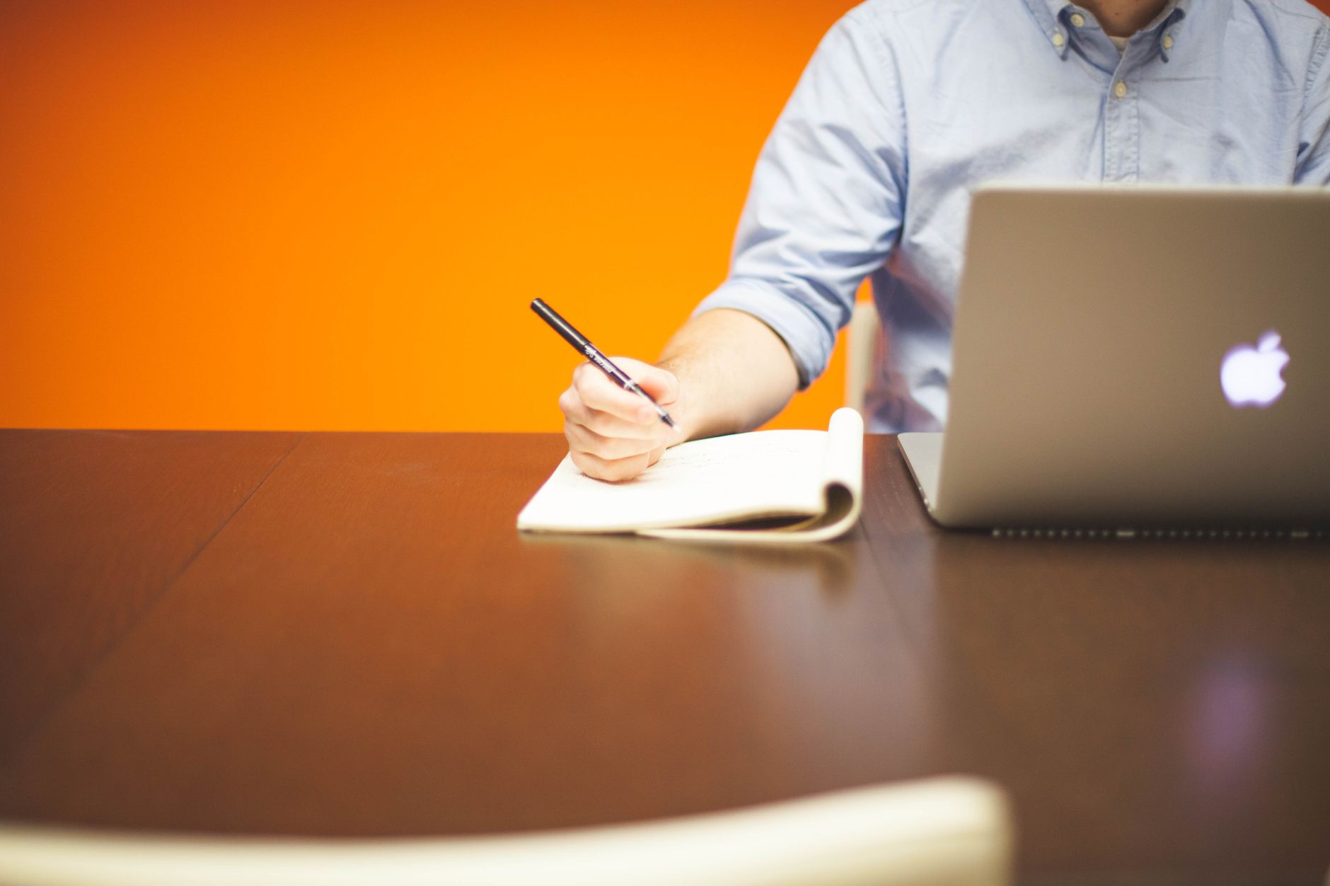 Volwassene schrijft notities in een notitieboek naast een laptop op een houten bureau met oranje achtergrond.