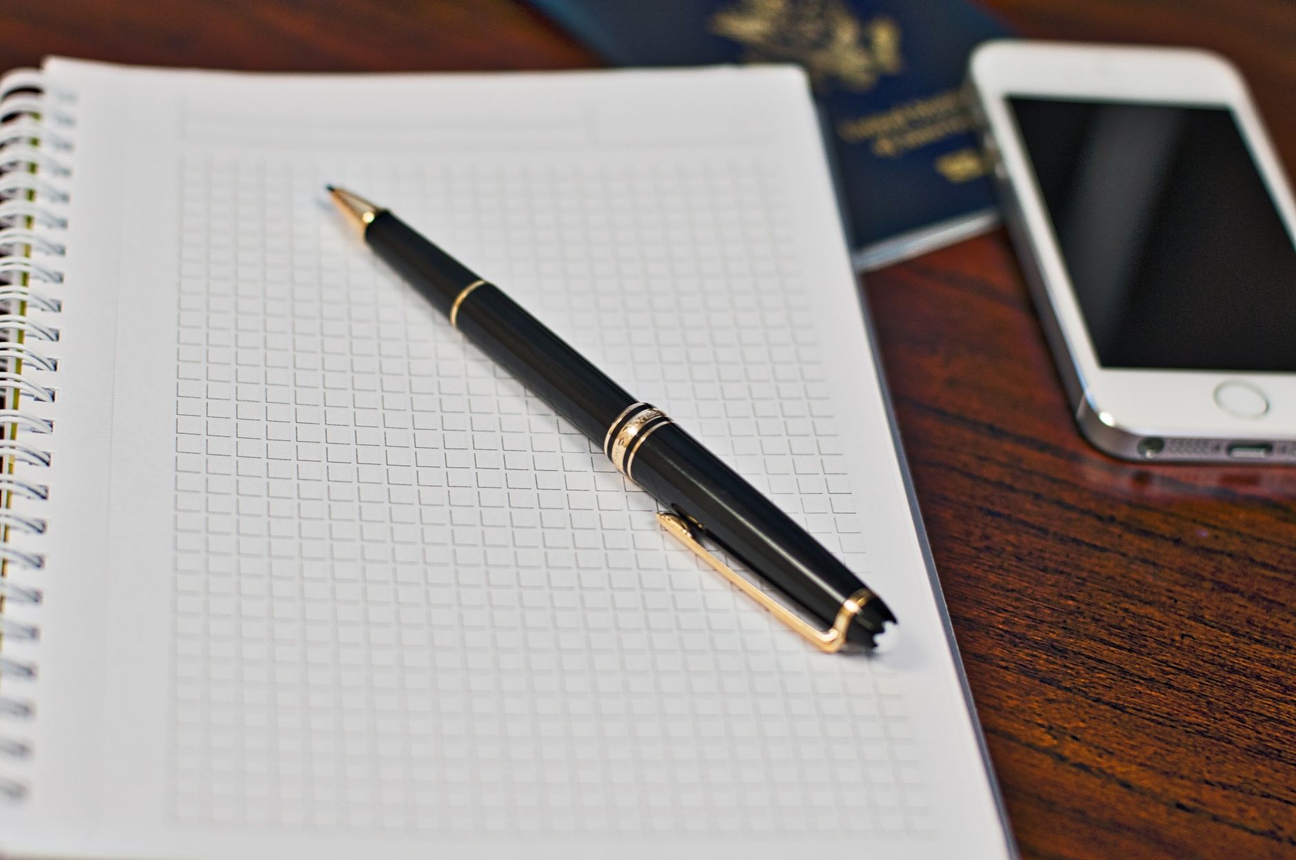 Zwarte pen op een geruit notitieboek met smartphone en paspoort op een houten bureau.